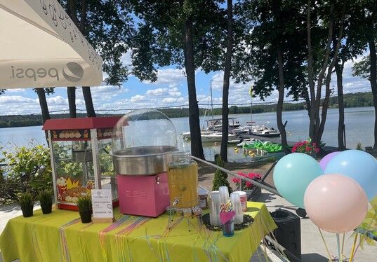 Lakeside snack station with popcorn and cotton‑candy machines, lemonade dispenser, pastel balloons and boat dock