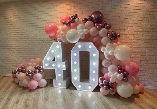 Illuminated '40' marquee numbers with a pink, white and rose-gold balloon arch against a white brick wall, party backdrop