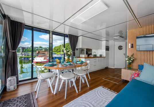Waterfront suite with floor-to-ceiling glass, marina view, set dining table, compact white kitchen, wooden accents and cozy lounge.
