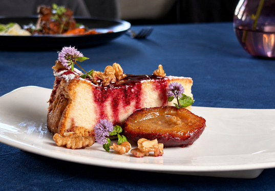 Käsekuchenstück mit Himbeer‑Glasur, karamellisierter Pflaume, Walnussstücken und essbaren violetten Blüten auf weißem Teller.