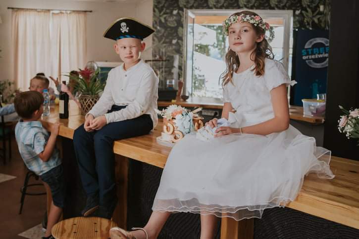 Girl in white dress with floral crown and boy in pirate hat sitting on a hotel wooden bar counter by a small celebration cake.
