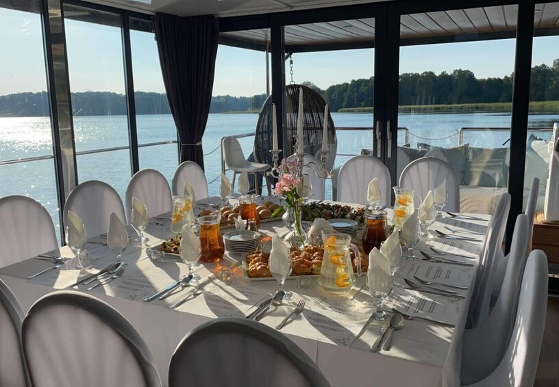 Glass-walled dining room on a boat with sunlit banquet table, white-covered chairs, pitchers with lemon, pastries, candelabra and tranquil lake view
