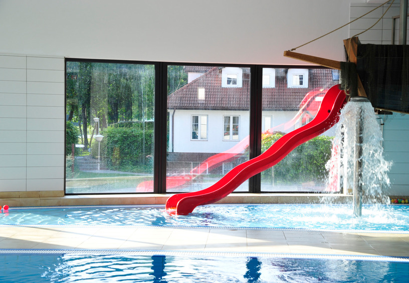 Indoor hotel pool with bright red twisting water slide, waterfall splash feature and large windows overlooking garden and neighboring building.