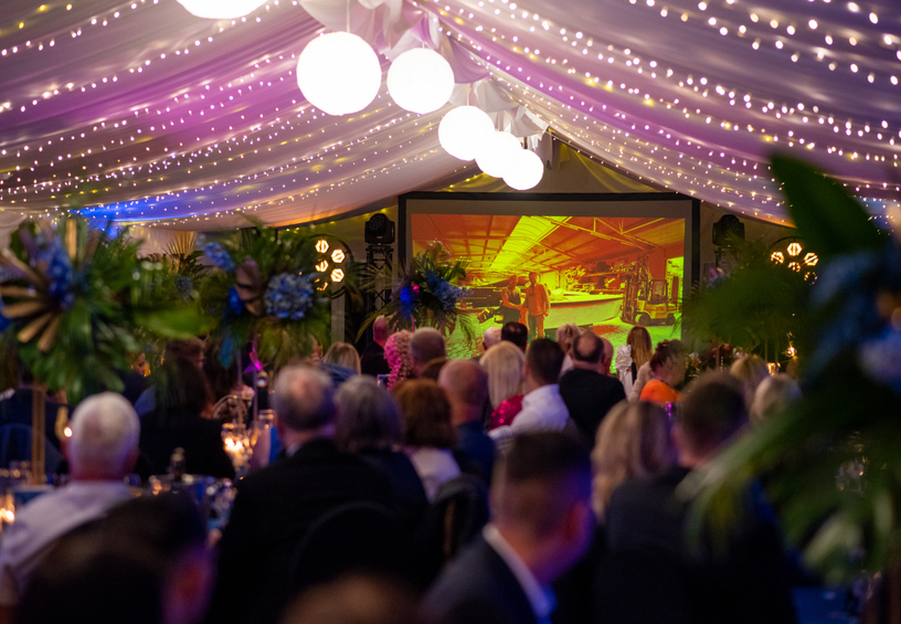 Banquet under a draped tent with string lights and round lanterns, seated guests, tall tropical centerpieces and a projection screen.