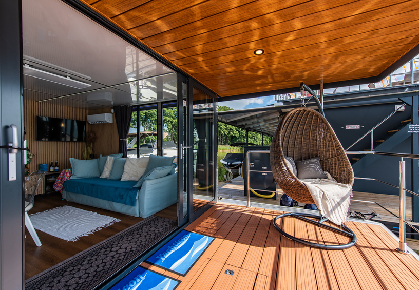 Private waterfront deck with wooden ceiling, hanging rattan chair and glass doors to a cozy lounge with sofa and TV.