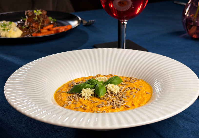 Cremiges Kürbisrisotto mit Sonnenblumenkernen, geriebenem Käse und Basilikum in weißer, gerillter Schale; Weinglas und Hauptgericht im Hintergrund.