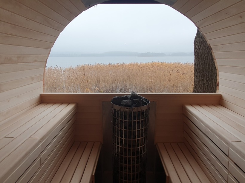Holzsauna am See mit gestuften Bänken, zentralem Ofen mit Saunasteinen und rundem Fenster auf Schilf und nebligen See