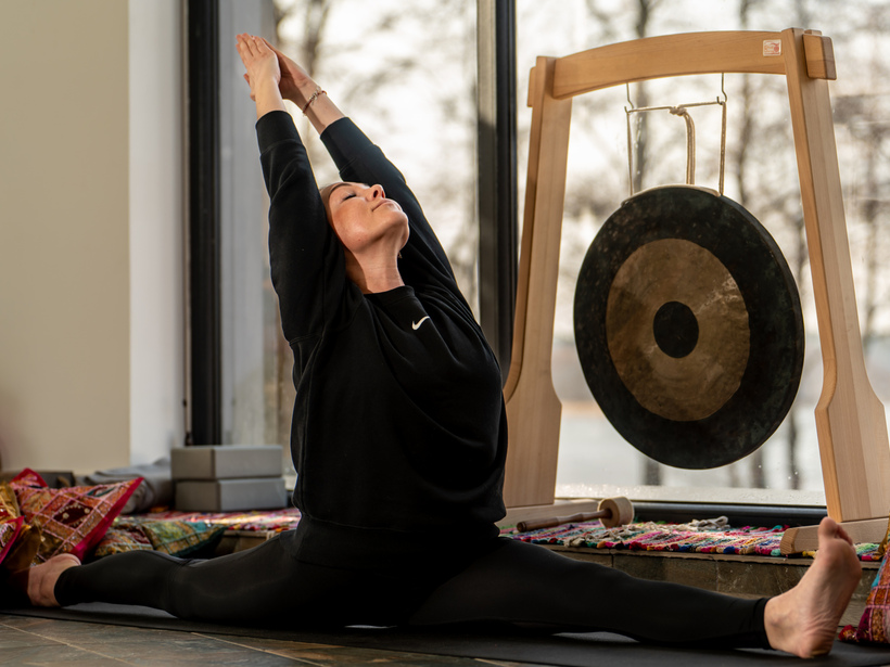Gość wykonuje szpagat i rozciąganie obok drewnianego gonga w jasnym studio wellness z kolorowymi poduszkami