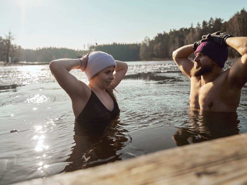 Couple taking a cold-water swim in a forest lake, wearing beanies and gloves, chest-deep near a wooden dock in soft morning light