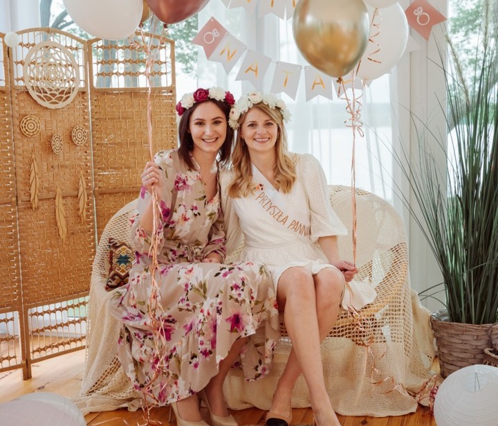 Panna młoda i przyjaciółka siedzą na wiklinowej sofie w jasnym salonie eventowym, wianki na głowach, balony, baner i pleciony dywan.