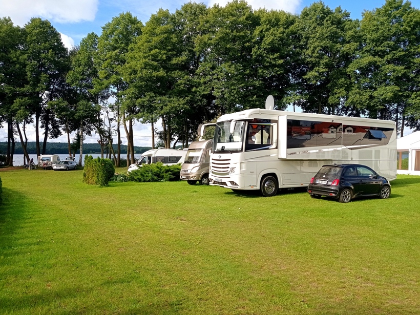 Grüner Campingplatz am See mit luxuriösen Reisemobilen und kleinem Auto unter hohen Bäumen, Gäste mit Fahrrädern am Ufer