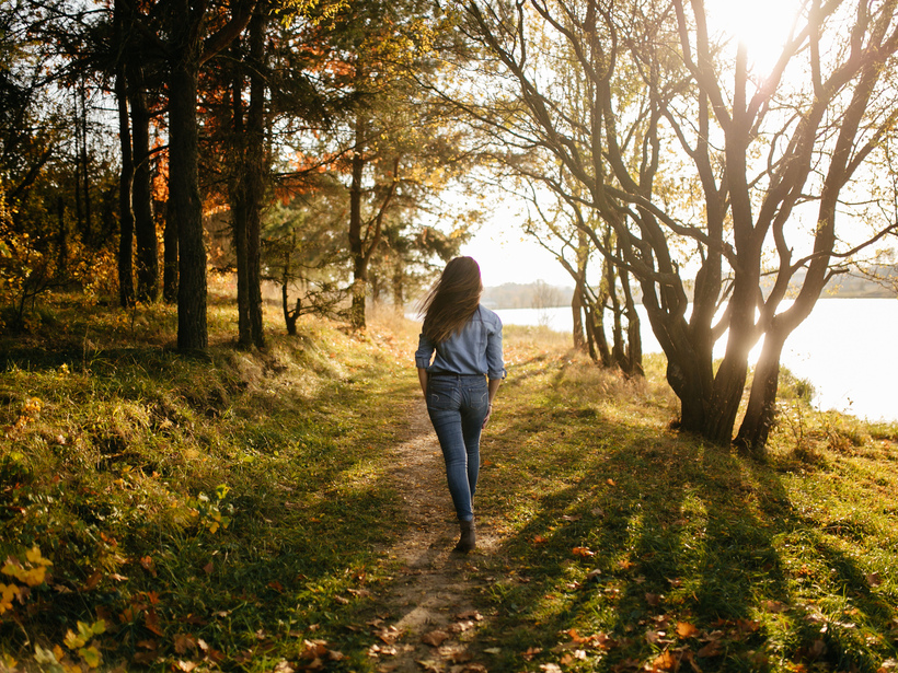 Woman strolling on a sunlit lakeside path through autumn trees, grassy trail and warm golden light.