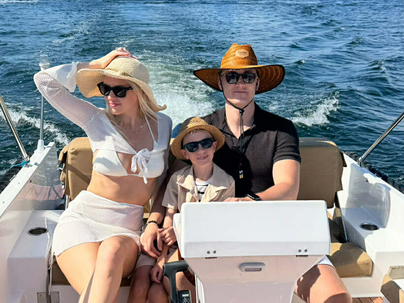 Familienbootfahrt auf dem See: Eltern und Kind mit Sonnenhüten und Sonnenbrillen am Steuer, Teakdeck, Heckwelle und bewaldetes Ufer.