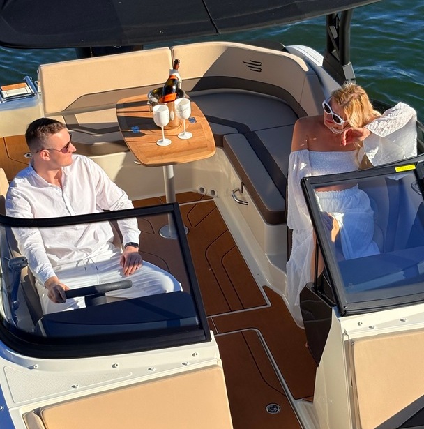 Paar auf privatem Motorboot — Mann am Steuer, Frau im weißen Kleid auf Teakdeck, Tisch mit Champagner und Gläsern