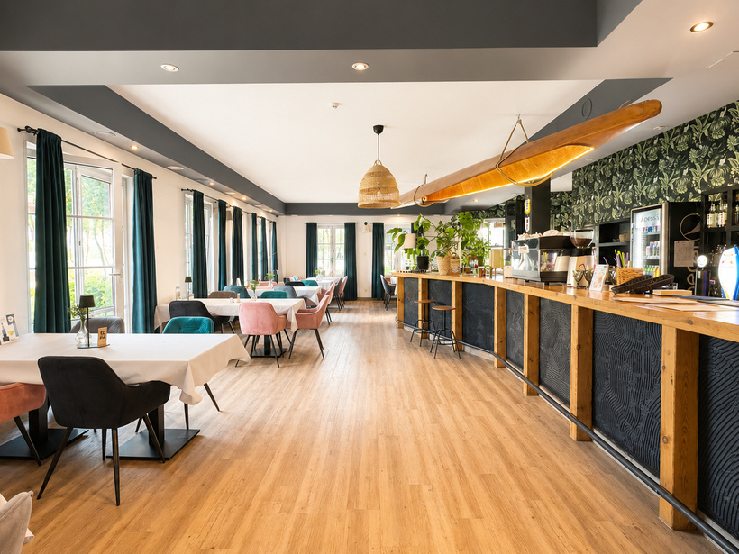 Helles Hotelrestaurant mit langer Holzbar, tropischer Tapete, türkisen und rosafarbenen Stühlen, großen Fenstern und Pflanzen.
