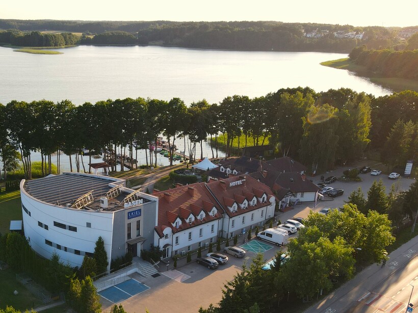 Hotel am See mit roten Ziegeldächern und modernem Spa-Gebäude, baumgesäumtem Ufer, Bootssteg und ruhigem Seeblick im Abendlicht