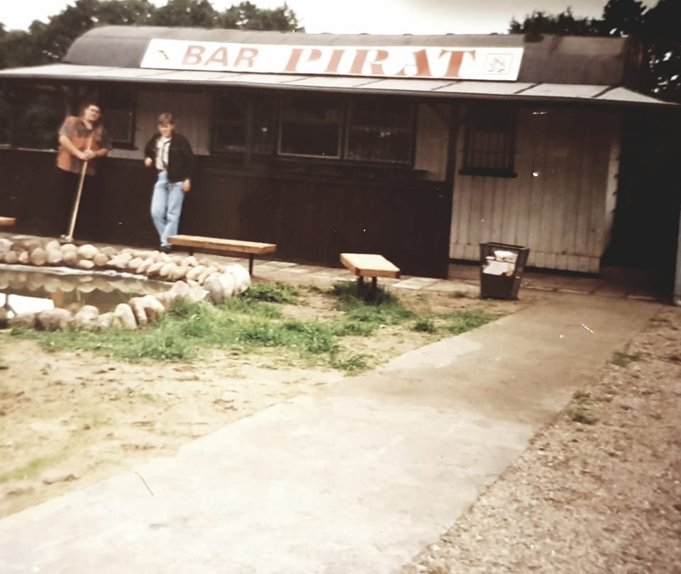 Rustic outdoor bar 'Pirat' with wooden counter, stone-edged pond, benches and two guests leaning by the bar