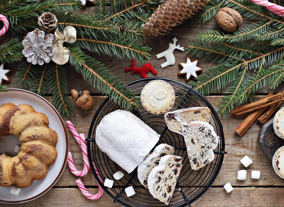 Festive dessert spread on wooden table: powdered stollen, bundt cake, mince pies, star cookies, candy canes and pine sprigs