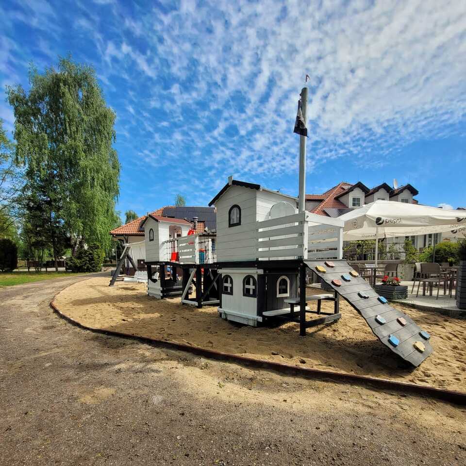 Hotel family playground with white wooden play-ship, climbing ramp over sand, terrace seating with umbrellas and red-tiled hotel facade