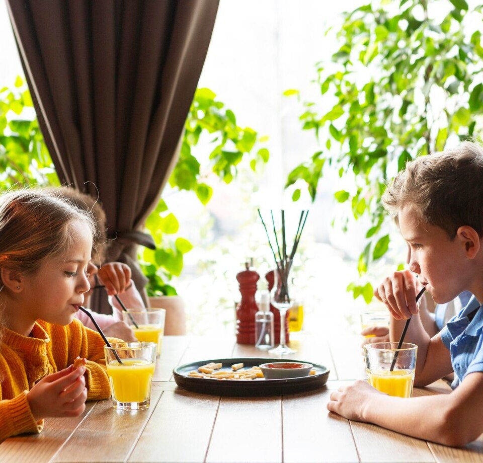 Two children sipping orange juice at a sunlit hotel breakfast table by the window, plate of pastries and green plants