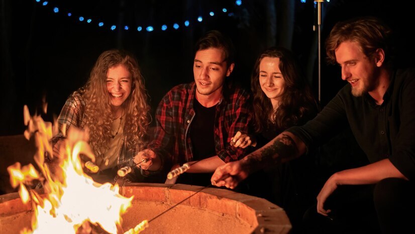 Freunde am Hotel-Feuerkorb im Freien bei Nacht, rösten Marshmallows unter Lichterketten in einer gemütlichen Lounge.