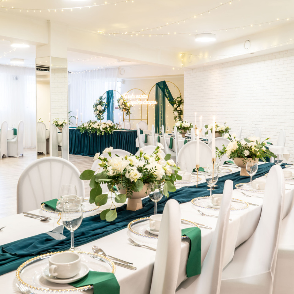 Elegancka sala bankietowa udekorowana na wesele: długie stoły z białymi pokrowcami, turkusowe bieżniki, kwiatowe kompozycje, łuk i lampki