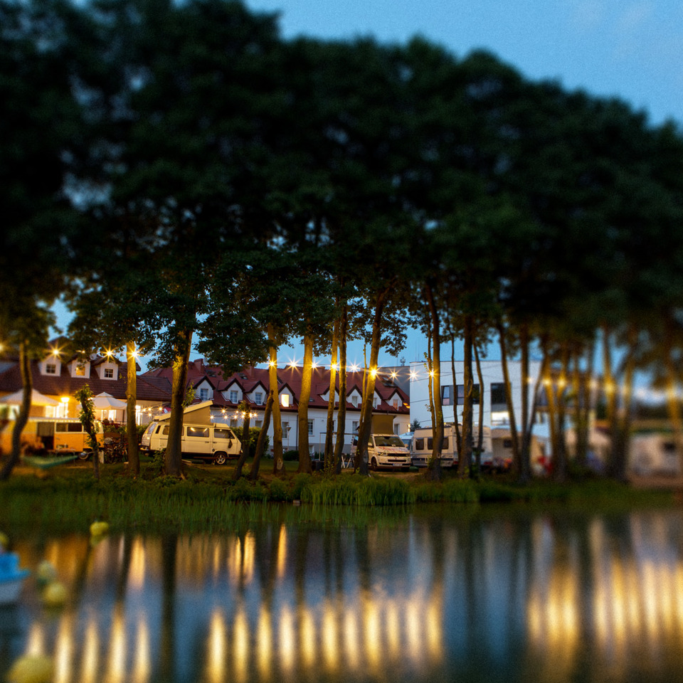 Lakeside guesthouse and camper area at dusk, string lights among tall trees reflecting on calm water, boats and red-roof building