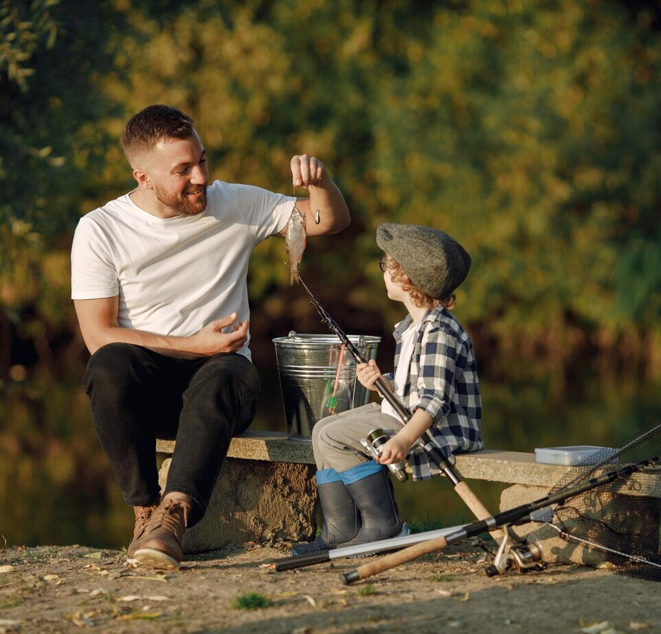 Father and son fishing from a lakeside bench at golden hour, small fish on the hook, fishing rods, bucket and landing net nearby