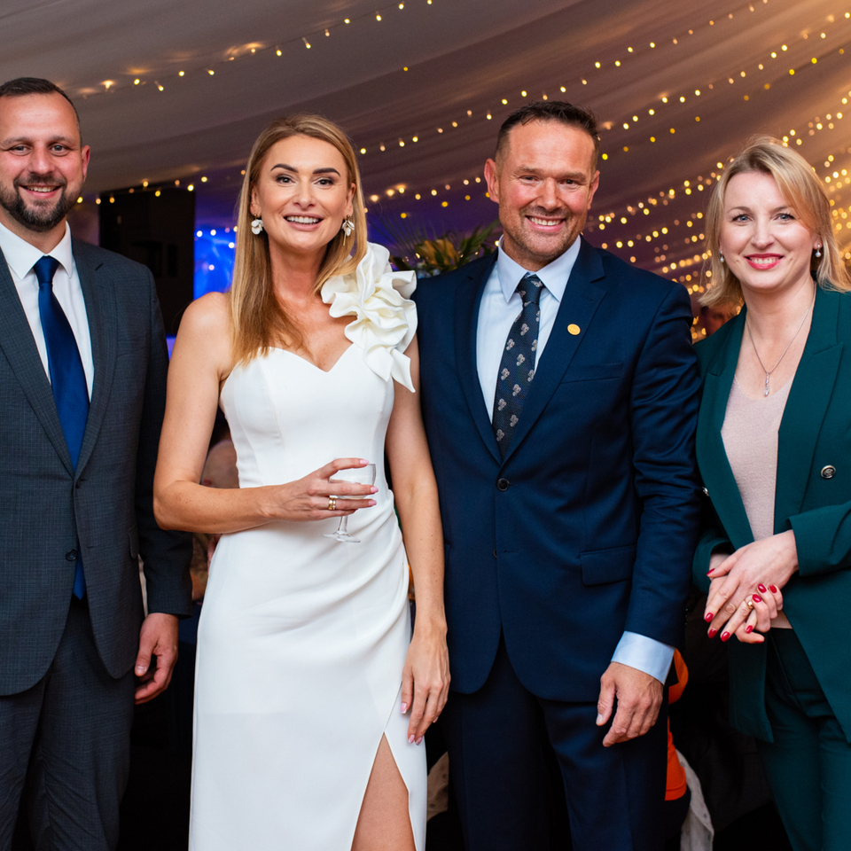 Four well-dressed guests smiling at an elegant hotel reception under fairy lights; woman in a white dress holding a wine glass.