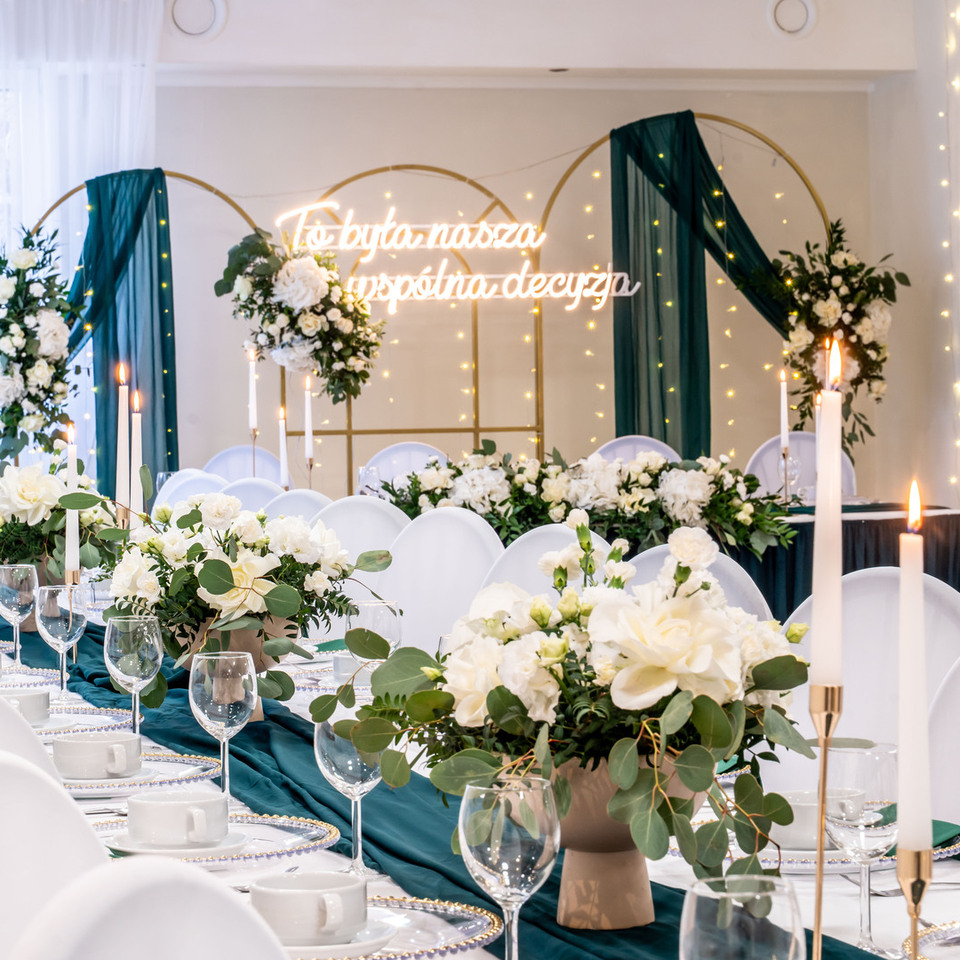 Elegant wedding banquet with table, white floral centerpieces, teal runner, lit candles, gold arches and fairy lights.