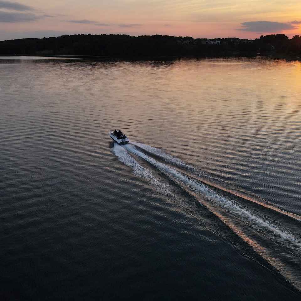 Kleines Motorboot durchquert ruhigen See bei Sonnenuntergang, goldene Spiegelungen und bewaldetes Ufer — perfekt für private Abendfahrten.