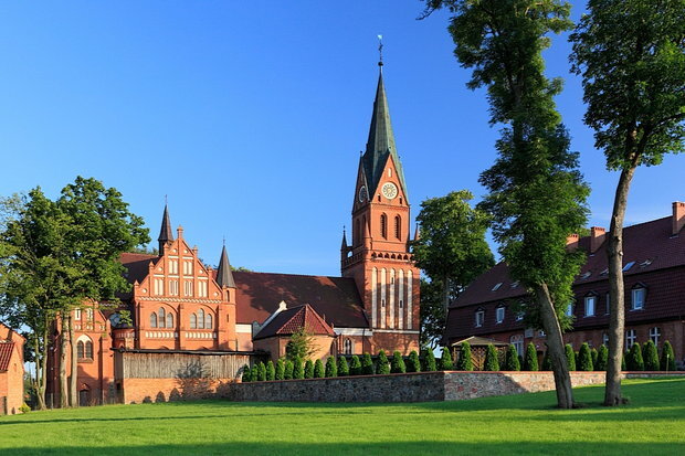 Historische Backsteinkirche mit hohem spitzem Turm neben gepflegtem Rasen und Bäumen unter klarem blauem Himmel