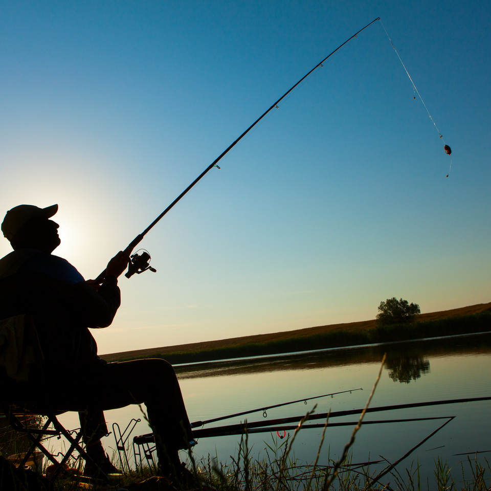 Silhouette of a person fishing from a folding chair at sunset by a calm lake with rod reflections