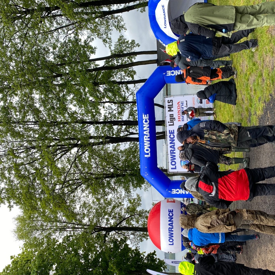 People in waterproof jackets gathered under a blue LOWRANCE arch at a rainy lakeside fishing event with tents and tall trees