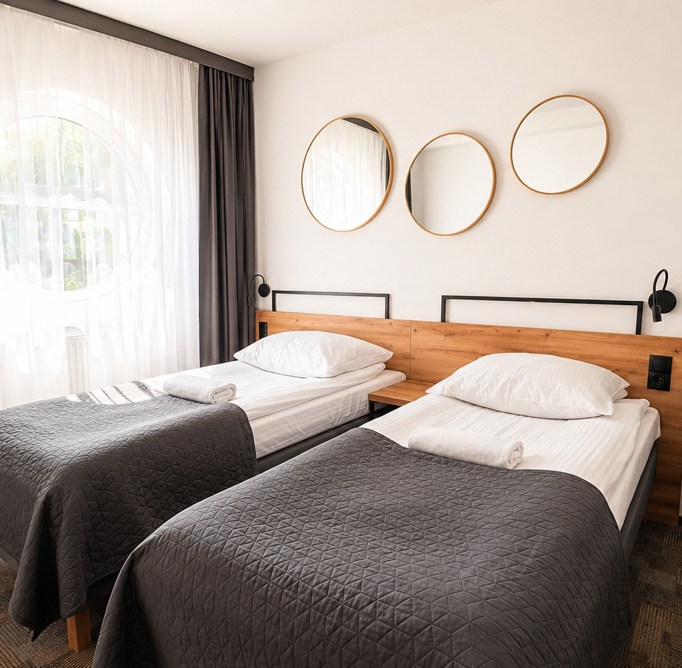 Zweibettzimmer mit zwei Einzelbetten, hölzernem Kopfteil, Leselampen und rundem Fenster mit transparentem Vorhang