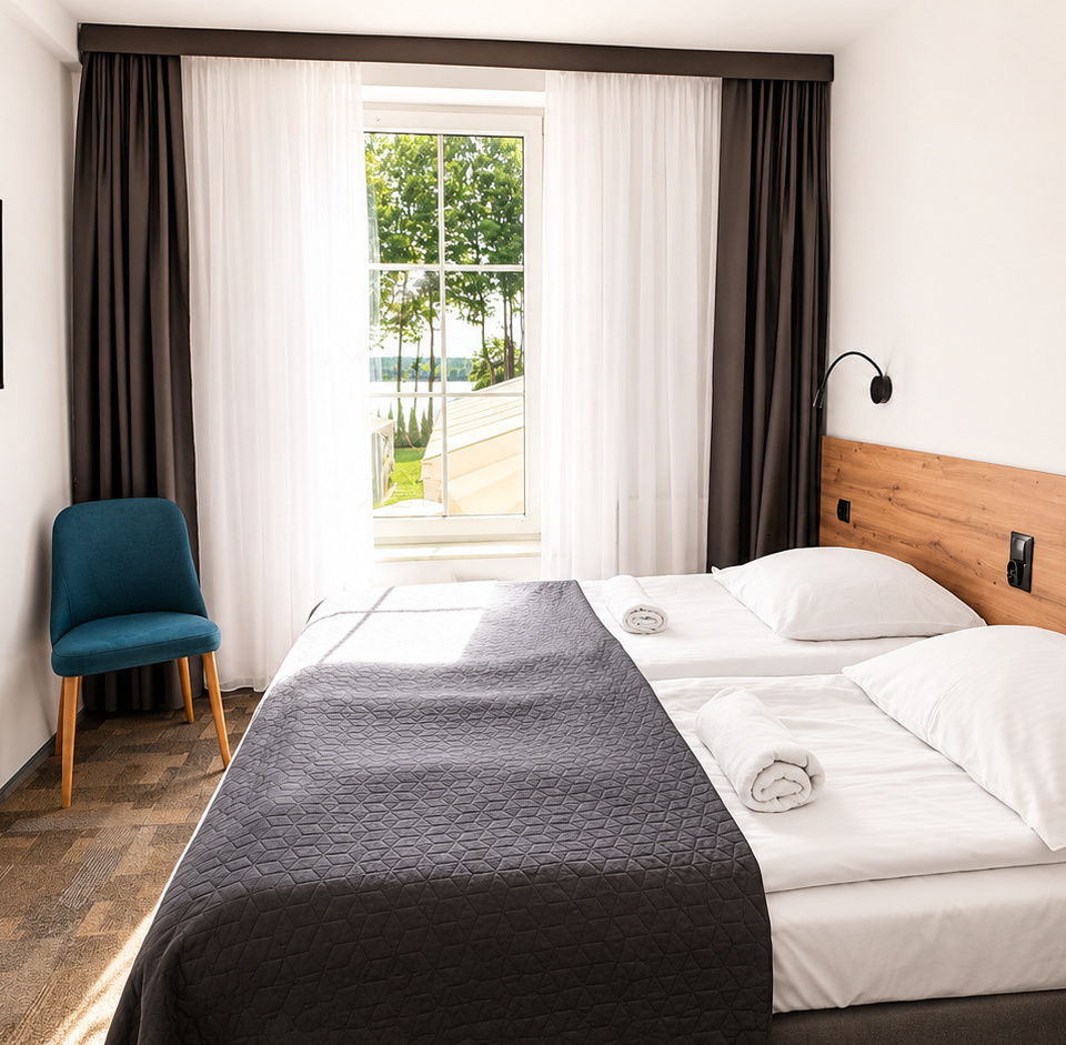 Helles, modernes Zweibettzimmer mit zwei Einzelbetten, Holzkopfteil, Wand-TV, türkisem Sessel, aufgerollten Handtüchern und Seeblick.