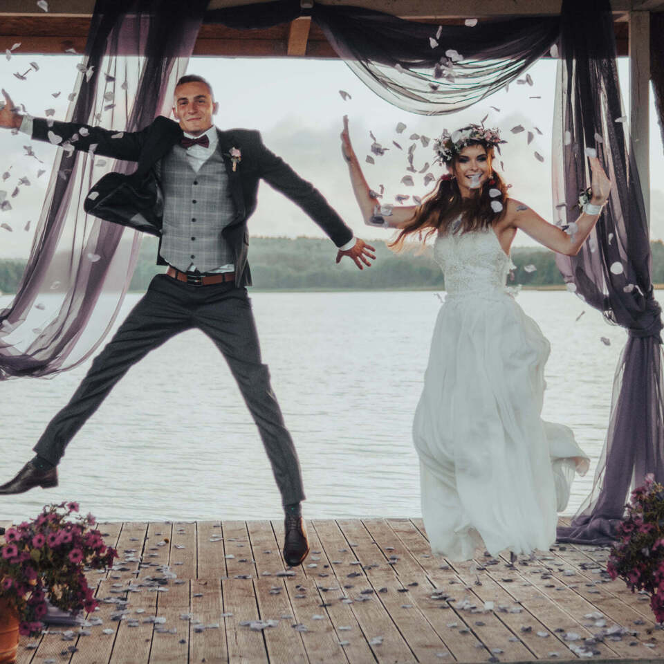Fröhliche Braut im Spitzenkleid mit Blumenkranz und Bräutigam springen unter Pavillon am See, Blütenblätter fallen.