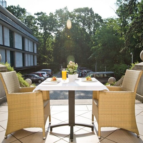 햇살 가득한 호텔 테라스, 2인용 식탁과 라탄 의자, 주스와 아침식사 접시, 녹음과 건물 전망.