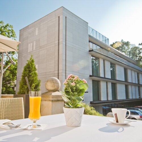 햇살 가득한 호텔 테라스의 테이블에 오렌지 주스, 커피 잔과 화분 꽃, 라탄 의자, 파라솔과 현대적 호텔 외관