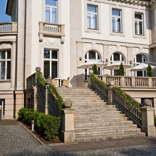 네오클래식 호텔 외관, 넓은 석조 계단이 테라스로 이어지고 발코니 난간과 파라솔이 있는 야외 식사 공간이 보임