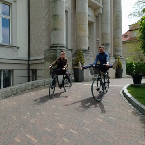 Dwójka gości radośnie jedzie hotelowymi rowerami przed neoklasycznym wejściem z wysokimi kamiennymi kolumnami.
