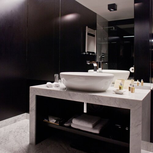 모던한 호텔 욕실, 대리석 세면대 위 흰색 볼형 세면기, 어메니티, 접힌 수건, 온열 타월걸이와 큰 거울.