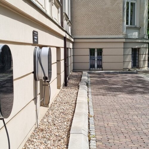 Dwa gniazda ładowania Porsche Destination na elewacji hotelu przy brukowanym podjeździe i zielonym żywopłocie