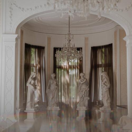 장식 아치와 크리스탈 샹들리를 갖춘 우아한 호텔 식당, 높은 창가의 대리석 석상 4점, 얇은 커튼과 흰 식탁보.