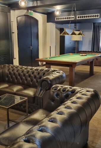 검은색 퀼팅 가죽 소파, 녹색 펠트 당구대와 황동 펜던트 조명이 있는 호텔 라운지형 아늑한 게임 공간.