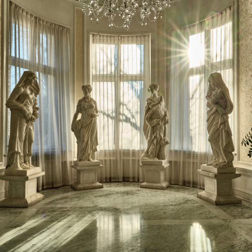 대리석 바닥과 크리스털 샹들리에, 햇빛이 드는 반원형 창가에 놓인 4점의 고전적 대리석 조각과 화분이 있는 고급 호텔 로비.