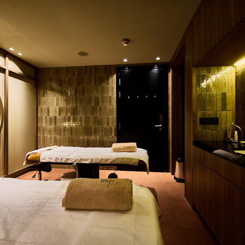 두 개의 마사지 테이블과 접힌 수건이 놓인 따뜻한 목재 인테리어 스파룸, 세면대와 은은한 조명.