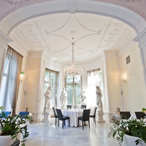 대리석 바닥과 크리스탈 샹들리에, 네 명용 원형 테이블, 고전 조각상과 높은 아치형 창이 있는 고급 호텔 다이닝룸