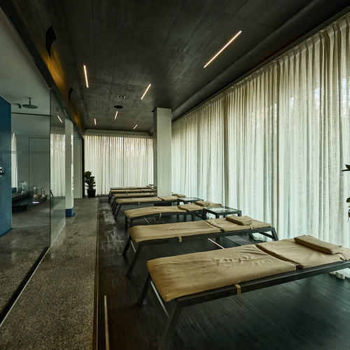 패드가 놓인 스파 라운지 일광욕용 의자, 천장까지 드리운 커튼, 은은한 조명과 파란 타일의 유리 습식 구역.