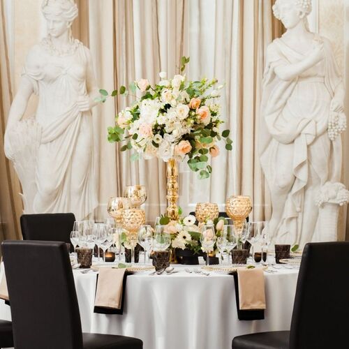 Elegancki okrągły stół na wykwintną kolację, złota kompozycja kwiatowa, kryształowe szkło, czarne krzesła i klasyczne rzeźby w tle